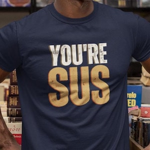 May include: A navy blue short-sleeved crew neck t-shirt featuring the text "YOU'RE SUS" printed in a distressed, bold, light-colored font across the chest. The shirt has a standard fit with a ribbed neckline and a solid dark blue fabric finish.