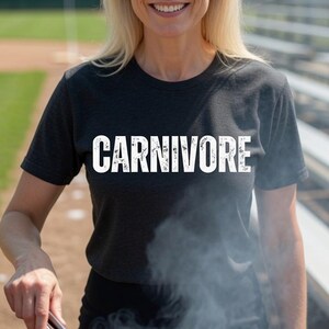 May include: A charcoal grey short-sleeved t-shirt featuring the word CARNIVORE printed in bold white capital letters across the chest. The shirt has a crew neckline and a standard fit, made from a heathered fabric texture.