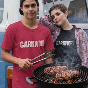 May include: A red t-shirt with the word CARNIVORE printed in white, a black t-shirt with the word CARNIVORE printed in white, a plaid button-down shirt, a black baseball cap, metal tongs, and a black charcoal grill with two grilled steaks.
