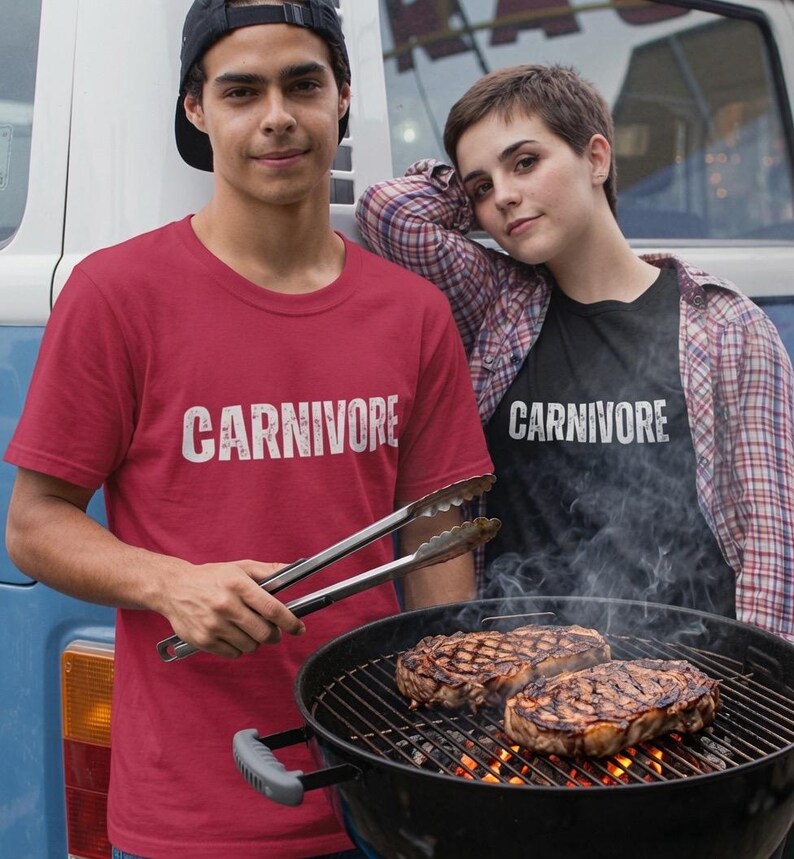 May include: A red t-shirt with the word CARNIVORE printed in white, a black t-shirt with the word CARNIVORE printed in white, a plaid button-down shirt, a black baseball cap, metal tongs, and a black charcoal grill with two grilled steaks.