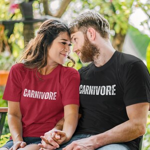 May include: A red short-sleeved t-shirt and a black short-sleeved t-shirt, both featuring the word CARNIVORE printed in a distressed white font across the chest. The shirts have a crew neckline and a standard fit, made of a solid cotton fabric.