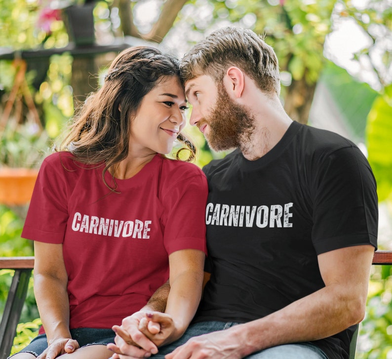 May include: A red short-sleeved t-shirt and a black short-sleeved t-shirt, both featuring the word CARNIVORE printed in a distressed white font across the chest. The shirts have a crew neckline and a standard fit, made of a solid cotton fabric.