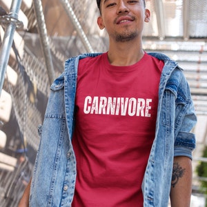 May include: A red short-sleeved t-shirt featuring the word CARNIVORE printed in white capital letters with a distressed texture across the chest, worn underneath a light blue denim jacket with metal buttons and visible stitching details.