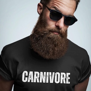 May include: A black short-sleeved t-shirt featuring the word CARNIVORE printed in white, distressed capital letters across the chest. The garment is worn by an individual with a thick brown beard and dark-rimmed sunglasses.