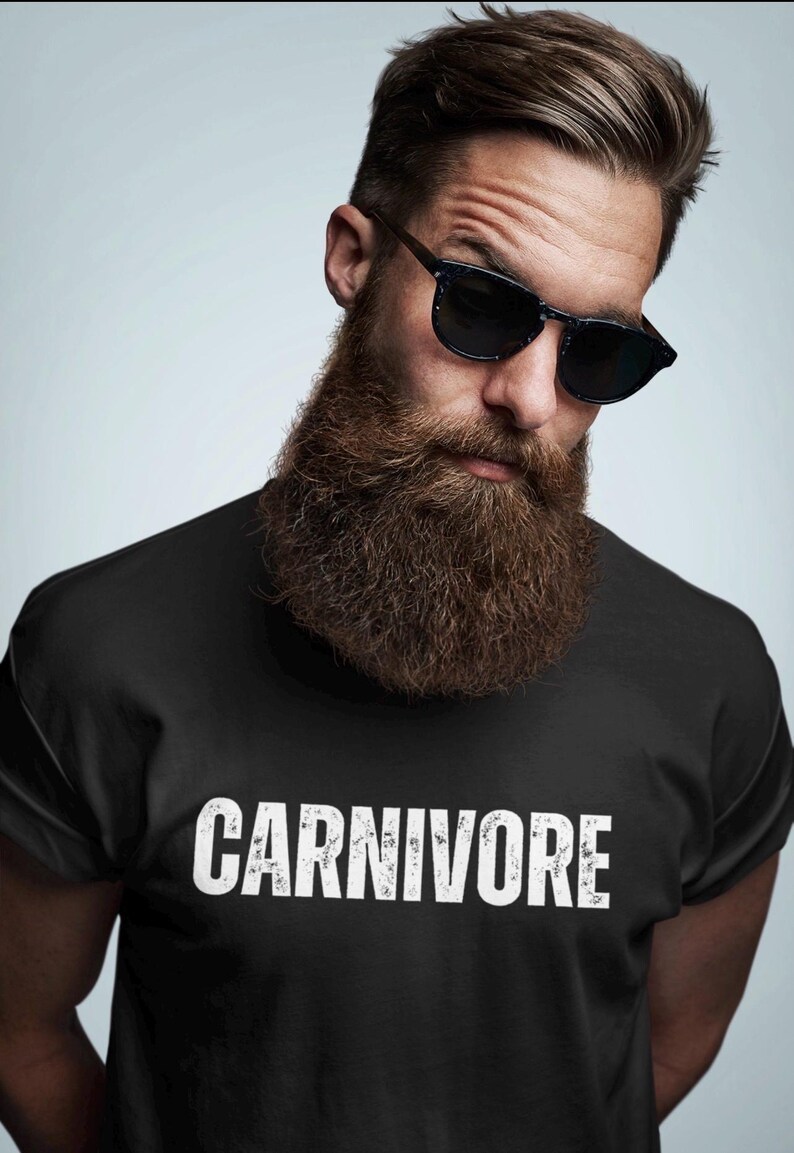 May include: A black short-sleeved t-shirt featuring the word CARNIVORE printed in white, distressed capital letters across the chest. The garment is worn by an individual with a thick brown beard and dark-rimmed sunglasses.