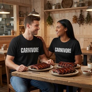 May include: Two black crew neck t-shirts featuring the word CARNIVORE printed in white distressed capital letters across the chest. The shirts are worn by two individuals sitting at a wooden table with plates of cooked ribs in front of them.