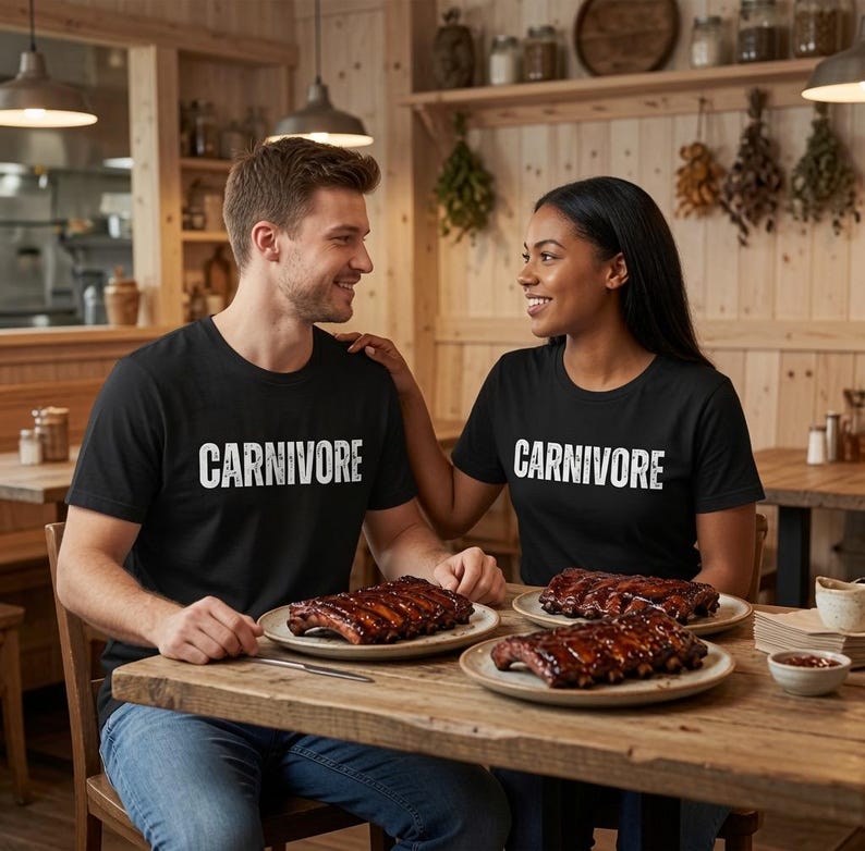 May include: Two black crew neck t-shirts featuring the word CARNIVORE printed in white distressed capital letters across the chest. The shirts are worn by two individuals sitting at a wooden table with plates of cooked ribs in front of them.