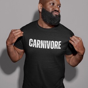 May include: A black short-sleeved t-shirt featuring the word CARNIVORE printed in a distressed white font across the chest. The garment has a standard crew neckline and a straight hem, providing a simple and direct design for casual wear.