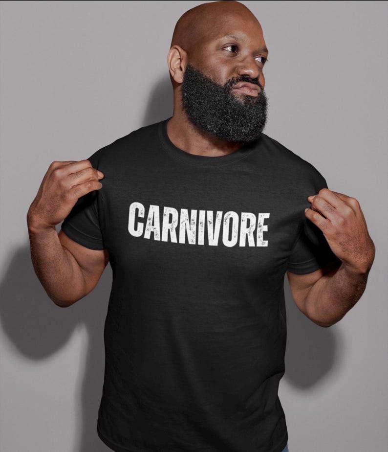 May include: A black short-sleeved t-shirt featuring the word CARNIVORE printed in a distressed white font across the chest. The garment has a standard crew neckline and a straight hem, providing a simple and direct design for casual wear.