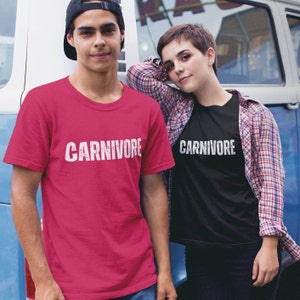 May include: A bright pink short-sleeved t-shirt and a black short-sleeved t-shirt, both featuring the word CARNIVORE printed in a distressed white font across the chest. The shirts have a crew neckline and a standard fit, showcasing a bold graphic design.
