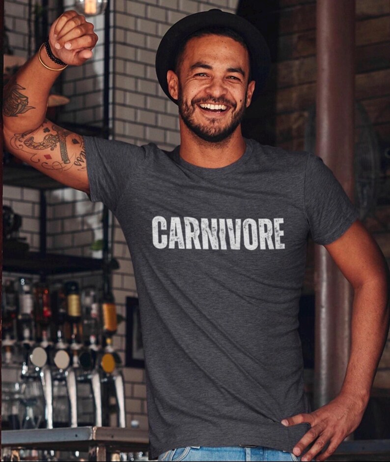 May include: A dark charcoal gray short-sleeved t-shirt featuring the word CARNIVORE printed in a distressed white font across the chest. The shirt has a crew neckline and a standard fit, constructed from a soft, heathered fabric material.