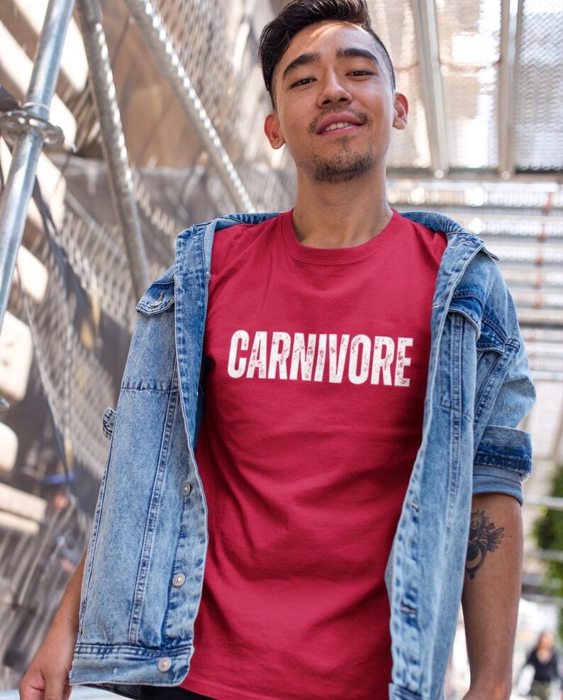 May include: A red short-sleeved t-shirt featuring the word CARNIVORE printed in white distressed capital letters across the chest, worn underneath a light blue denim jacket with silver buttons and a visible chest pocket detail.