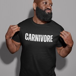 May include: A black short-sleeved t-shirt featuring the word CARNIVORE printed in a distressed white font across the chest. The garment has a classic crew neckline and a standard fit, showcasing a bold graphic design on the front of the shirt.