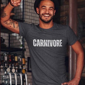 May include: A dark charcoal gray short-sleeved t-shirt featuring the word CARNIVORE printed in a distressed white font across the chest. The shirt has a crew neckline and a heathered texture, providing a casual look for everyday wear.