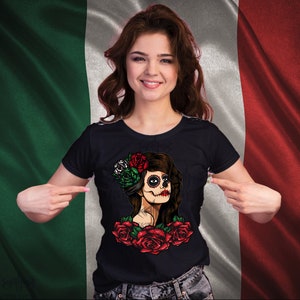 May include: A black t-shirt with a colorful illustration of a woman with a sugar skull face and flowers. The woman is wearing a flower crown with red, white, and green flowers. The background is a blurry image of a Mexican flag.