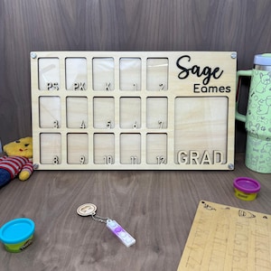 May include: A wooden board with 12 rectangular slots for photos, labeled with numbers from PS to 12. The board has the name "Sage Eames" and the word "GRAD" printed on it.