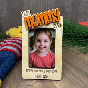 May include: A wooden photo frame with the words "Best Mommy Ever" in orange lettering. The frame has a photo of a young girl smiling. The text below the photo reads "Happy Mother's Day 2025 Love, Evie".