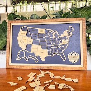 May include: Wooden United States map puzzle with individual state pieces. The map is framed in a light brown wood and set against a dark blue background. The puzzle includes the text "DANIEL & CARA'S" and is displayed on a wooden surface.