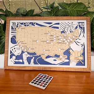 May include: Wooden baseball stadium map of the United States, with stadium locations marked. The map is framed in a brown wooden frame, with a blue and white background. A small remote with colored dots is in front of the map.