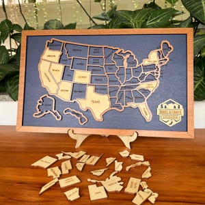 May include: A wooden United States map puzzle with individual state pieces. The map is framed in a light brown wood, with a dark blue background. The state names are engraved. The puzzle includes a stand and loose state pieces.