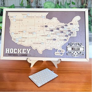 May include: A wooden hockey-themed wall art piece featuring a map of the United States with hockey puck icons marking locations. The words "HOCKEY NATIONAL LEAGUE" are displayed. A smaller wooden piece with hockey puck icons is in front of the art.