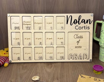 May include: A wooden photo frame with 12 slots for pictures, labeled with grades from preschool to 12th grade. The frame is personalized with the name "Nolan Cortis" and the text "Class of 2039 GRAD".