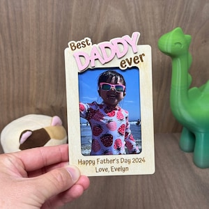 May include: A wooden photo frame with the words "Best Daddy ever" in pink letters. The frame has a photo of a young girl wearing sunglasses and a strawberry print shirt. The text below the photo reads "Happy Father's Day 2024 Love, Evelyn."