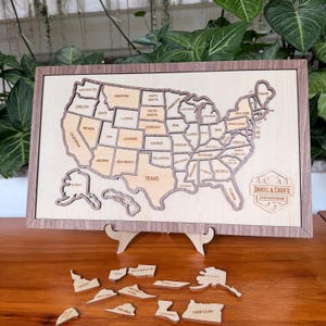 Peut inclure: Puzzle en bois représentant une carte des États-Unis, avec des pièces individuelles pour chaque État. La carte est encadrée dans un bois brun clair et comprend le texte "DANIEL & CARA'S ADVENTURE MAP".