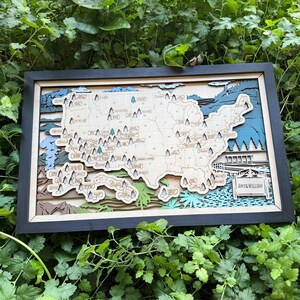 May include: A framed, layered wooden map of the United States, with a black frame. The map is intricately detailed with various colours and shapes, including mountains, trees, and water features. The sign at the bottom reads "AMY & WILLIAM".