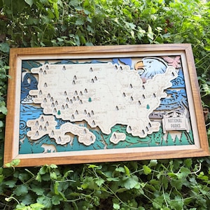 May include: Wooden layered map of the United States with a detailed design. The map features a brown wooden frame, with a light-colored map of the US, and a blue, green, and white background. The words "NATIONAL PARKS" are visible.