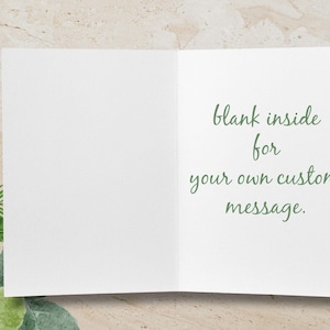 Pode incluir: Um cart&atilde;o de felicita&ccedil;&otilde;es branco aberto com o texto "blank inside for your own custom message" em verde. O fundo &eacute; bege claro com folhagem verde e branca no canto.