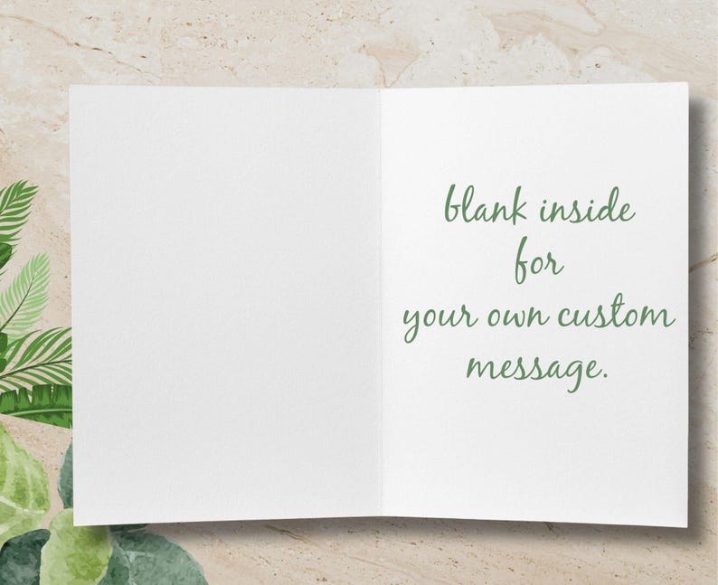 Pode incluir: Um cart&atilde;o de felicita&ccedil;&otilde;es branco aberto com o texto "blank inside for your own custom message" em verde. O fundo &eacute; bege claro com folhagem verde e branca no canto.