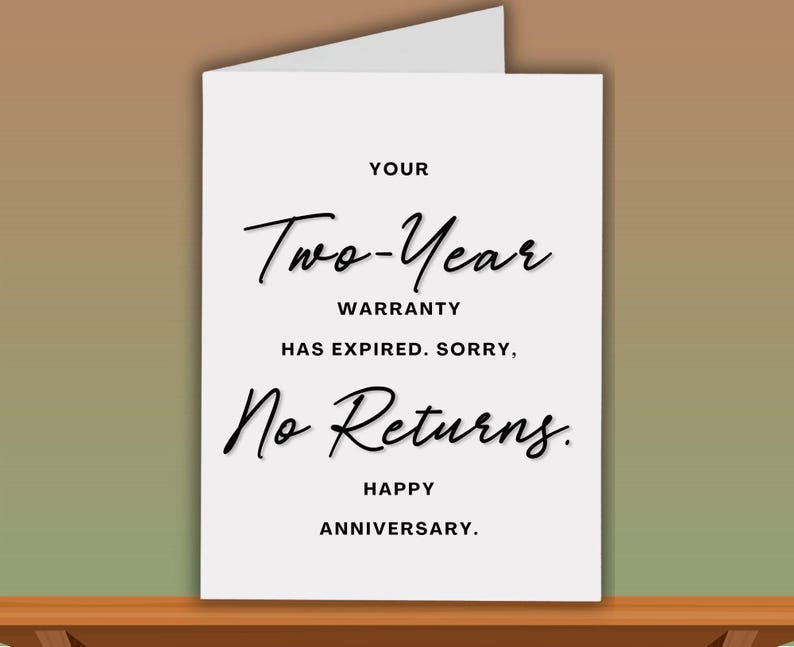 Pode incluir: Cart&atilde;o branco com texto preto: "YOUR Two-Year WARRANTY HAS EXPIRED. SORRY, No Returns. HAPPY ANNIVERSARY." O cart&atilde;o est&aacute; sobre uma superf&iacute;cie de madeira.