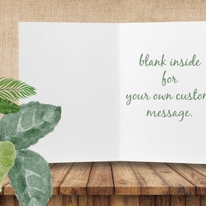 Op de afbeelding: Een open, blanco witte wenskaart met de tekst "blank inside for your own custom message" in groene letters. De kaart ligt op een houten oppervlak, met groene bladeren. Ideaal voor een persoonlijke boodschap.