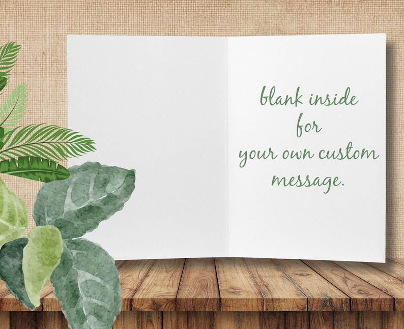 Op de afbeelding: Een open, blanco witte wenskaart met de tekst "blank inside for your own custom message" in groene letters. De kaart ligt op een houten oppervlak, met groene bladeren. Ideaal voor een persoonlijke boodschap.