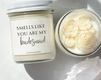 Może przedstawiać: Biała świeca z etykietą z napisem "Smells like you are my bridesmaid" i nazwą marki "Tansagamore". Świeca znajduje się w przezroczystym szklanym słoiku z białą pokrywką. W tle znajdują się białe kwiaty.