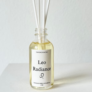Puede incluir: Una botella difusora de caña de vidrio transparente llena de líquido amarillo y cañas blancas. La etiqueta dice "Leo Radiance" con el símbolo del zodiaco Leo y "Aceite difusor de caña hecho a mano 2 oz."
