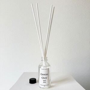 May include: A clear glass aromatherapy diffuser bottle with white reeds. The label reads "Gemini Glow" with the Gemini zodiac symbol. A black cap sits beside the bottle. The diffuser is on a white surface.