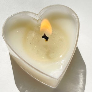 May include: A heart-shaped candle with a clear glass container and a single wick burning with a bright yellow flame.
