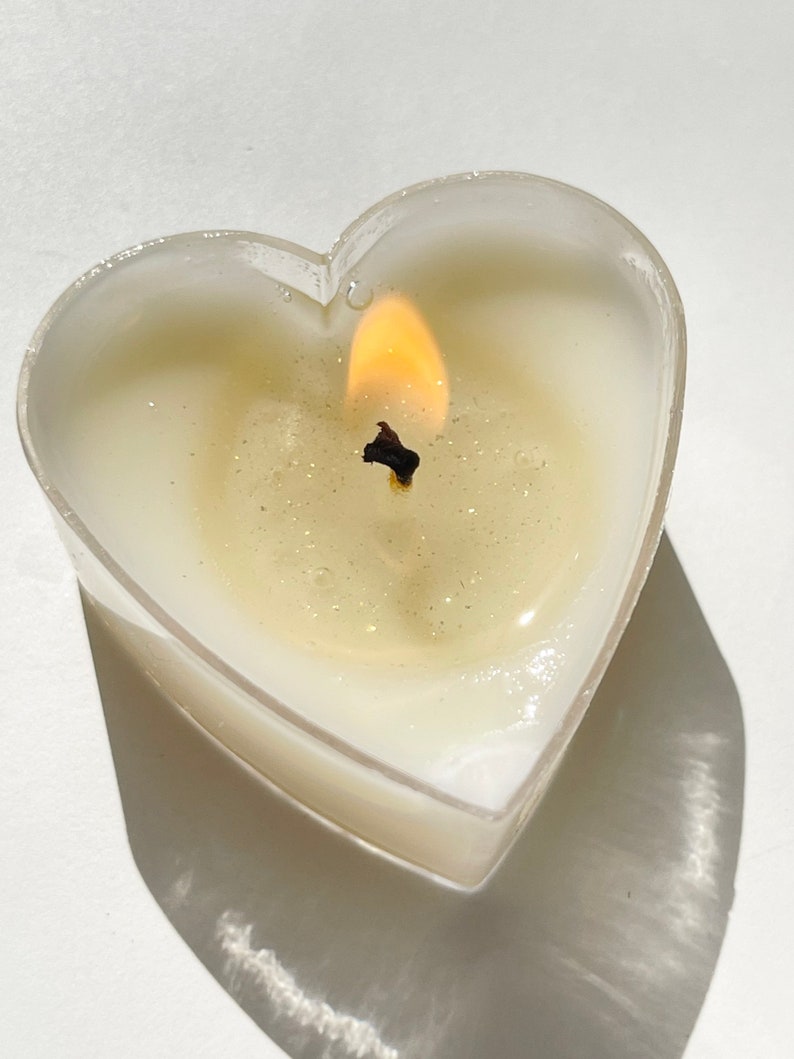May include: A heart-shaped candle with a clear glass container and a single wick burning with a bright yellow flame.