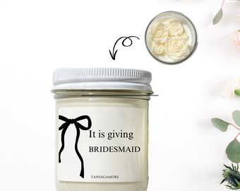 Op de afbeelding: Een witte kaars in een glazen pot met een wit deksel. De kaars heeft een wit etiket met zwarte tekst die "It is giving BRIDESMAID" en een zwarte strik tekening leest. De kaars is omringd door witte bloemen.