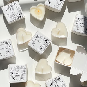 May include: Assortment of heart-shaped candles in white boxes with floral designs and black text. The candle scents include: Coconut Santal, Strawberry, Rose Garden, After Rain, Fairy, The Fairy Forest, Daisy, Wonder Land.