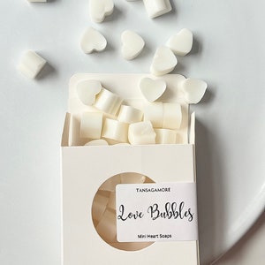Op de afbeelding: Een witte doos met een rond venster dat witte hartvormige zeepjes laat zien. De doos heeft een label met de tekst "Love Bubbles" en "Mini Heart Soaps".