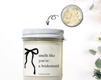 Op de afbeelding: Een witte kaars in een glazen pot met een wit deksel. De kaars heeft een zwart lint eromheen gebonden en een etiket met de tekst "smells like you're a bridesmaid" en "TANSAGAMORE".