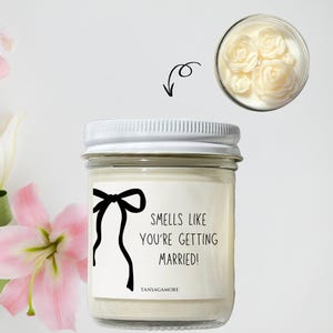 May include: White candle in a glass jar with a white lid. The candle has a label that says "Smells like you're getting married!" with a black bow drawn on the label. The candle is on a white background.