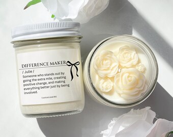 Op de afbeelding: Een witte kaars met een label waarop staat "Difference Maker / Julie / Iemand die opvalt door die extra stap te zetten, positieve verandering te creëren en alles gewoonweg te verbeteren door betrokken te zijn. TANSAGAMORE". De kaars is versierd met witte wasrozen.