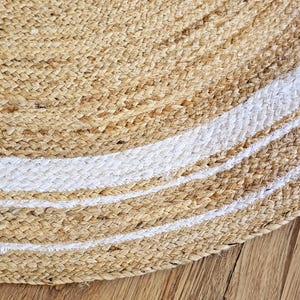 Peut inclure: Gros plan sur un tapis rond en jute de couleur beige naturel avec des rayures blanches. Le tapis est fait de fibres tiss&eacute;es et a une surface textur&eacute;e. Le tapis est sur un plancher en bois.