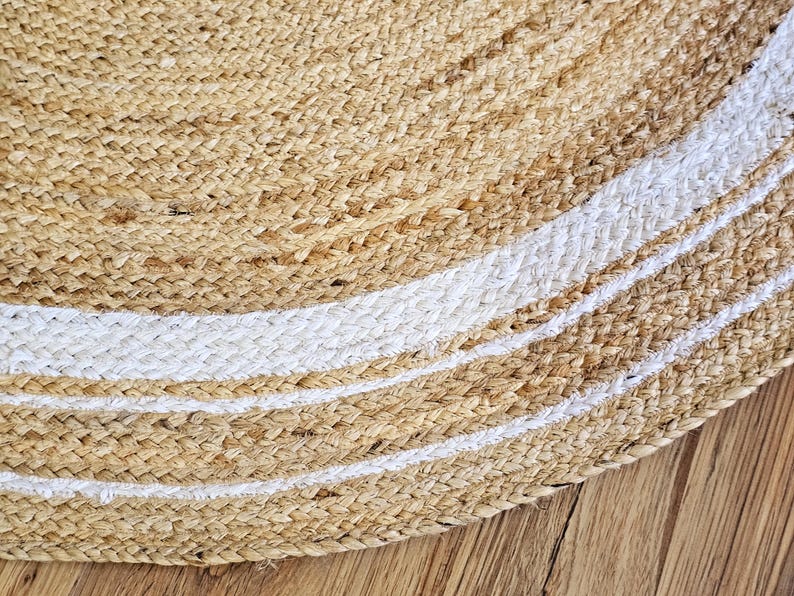 Peut inclure: Gros plan sur un tapis rond en jute de couleur beige naturel avec des rayures blanches. Le tapis est fait de fibres tiss&eacute;es et a une surface textur&eacute;e. Le tapis est sur un plancher en bois.
