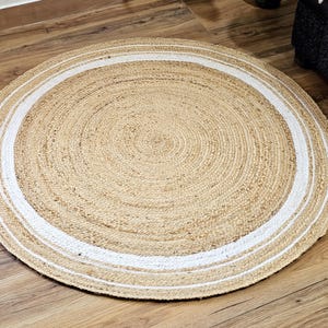 Peut inclure: Un tapis rond en jute de couleur beige naturel avec une bordure blanche. Le tapis est fait de fibres tiss&eacute;es et est pos&eacute; sur un sol en bois. Le diam&egrave;tre du tapis est d'environ 120 cm.