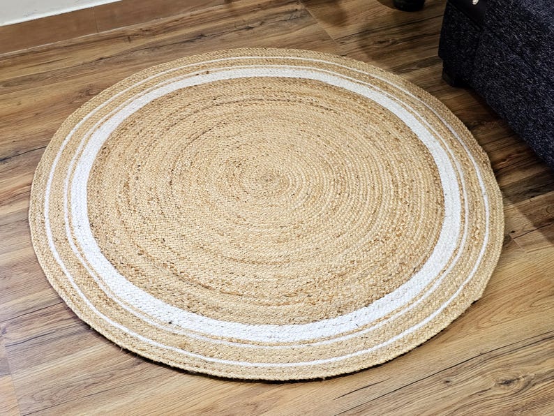 Peut inclure: Un tapis rond en jute de couleur beige naturel avec une bordure blanche. Le tapis est fait de fibres tiss&eacute;es et est pos&eacute; sur un sol en bois. Le diam&egrave;tre du tapis est d'environ 120 cm.